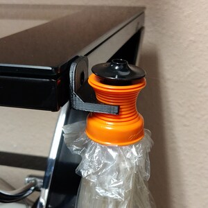 Op de afbeelding: Een 3D-geprinte oranje en zwarte plastic tasjeshouder bevestigd aan een zwart oppervlak. De houder heeft een kleine, ronde, zwarte bovenkant en een geribbeld, oranje lichaam. Een doorzichtige plastic tas hangt aan de houder.