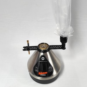 May include: A silver Volcano Hybrid vaporizer with a black base and a clear plastic bag attached to the top. The vaporizer has a digital display with the word "Volcano" and the word "Hybrid" printed on it.