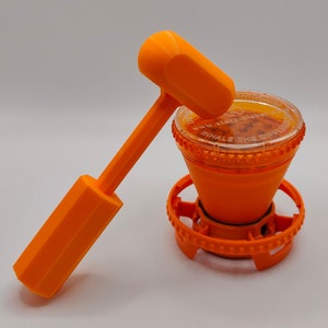 May include: An orange plastic grinder with a clear lid and a separate orange plastic mallet. The grinder has a base with a gear-like design and the text "INHALE THE DIFFERENCE" on the lid.