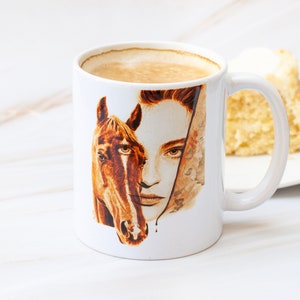 May include: White ceramic mug with a brown and white illustration of a woman's face and a horse's head. The mug is filled with coffee.