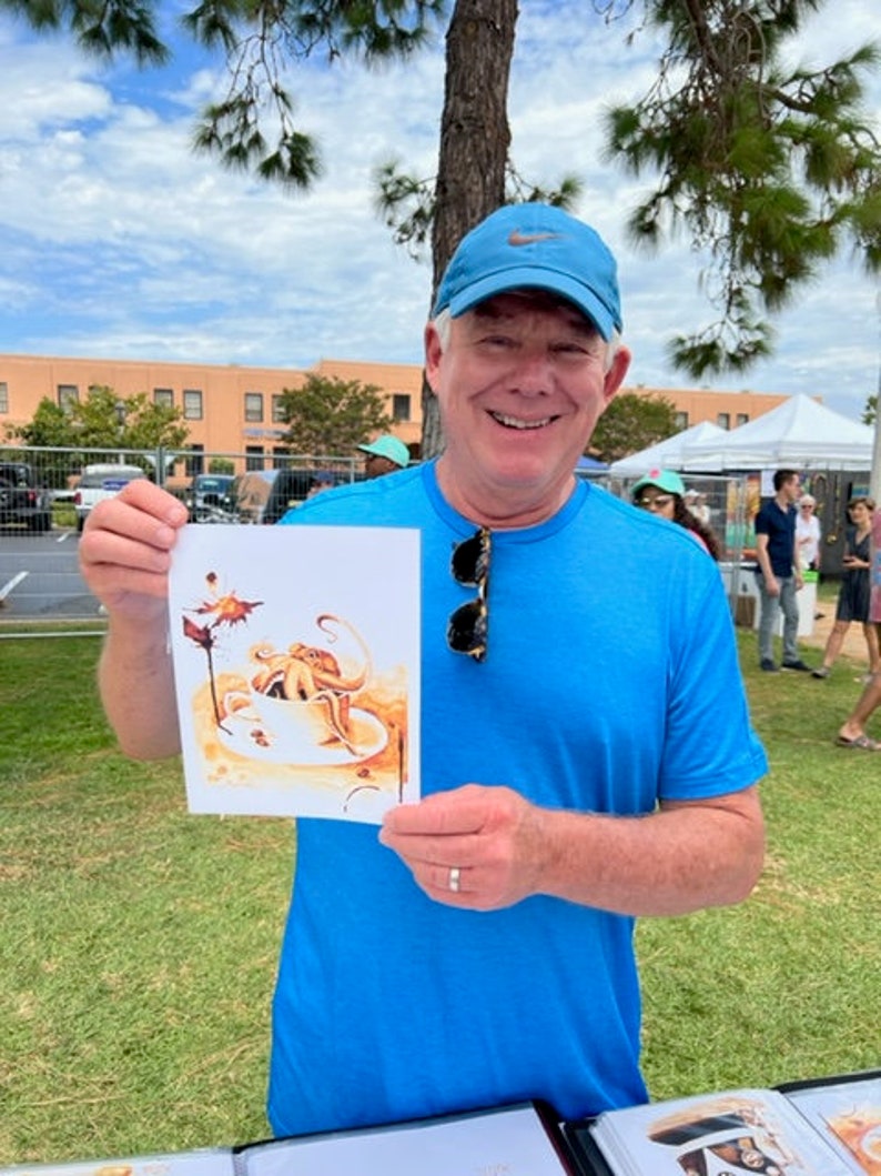 Puede incluir: Un hombre con una camisa azul y una gorra de b&eacute;isbol azul sostiene una tarjeta blanca con una ilustraci&oacute;n de acuarela de un pulpo en una taza de caf&eacute;. La ilustraci&oacute;n est&aacute; en tonos marrones y naranjas.