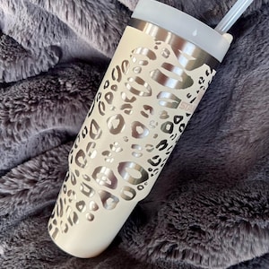 Puede incluir: Un vaso de color crema con un dise&ntilde;o de estampado de leopardo plateado. El vaso tiene un borde plateado, una tapa transparente y una pajita transparente. El vaso est&aacute; sobre una manta gris y esponjosa. La palabra "STAR" es visible.