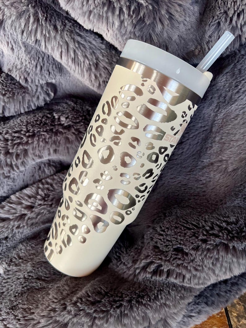 Puede incluir: Un vaso de color crema con un dise&ntilde;o de estampado de leopardo plateado. El vaso tiene un borde plateado, una tapa transparente y una pajita transparente. El vaso est&aacute; sobre una manta gris y esponjosa. La palabra "STAR" es visible.