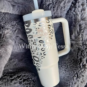 Puede incluir: Un vaso Stanley blanco con asa y tapa, con un dise&ntilde;o de estampado de leopardo plateado. El vaso tiene una pajita y la marca "STANLEY" es visible. El texto "With Carey by Tessa" tambi&eacute;n es visible.