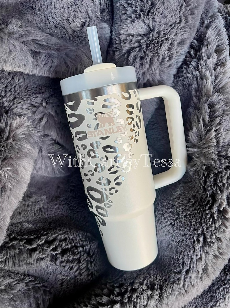 Puede incluir: Un vaso Stanley blanco con asa y tapa, con un dise&ntilde;o de estampado de leopardo plateado. El vaso tiene una pajita y la marca "STANLEY" es visible. El texto "With Carey by Tessa" tambi&eacute;n es visible.