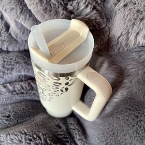 Puede incluir: Taza de viaje color crema con un dise&ntilde;o de estampado de leopardo plateado. La taza tiene un asa, una tapa con pajita y un cierre abatible color crema. La taza est&aacute; sobre una manta gris y esponjosa.