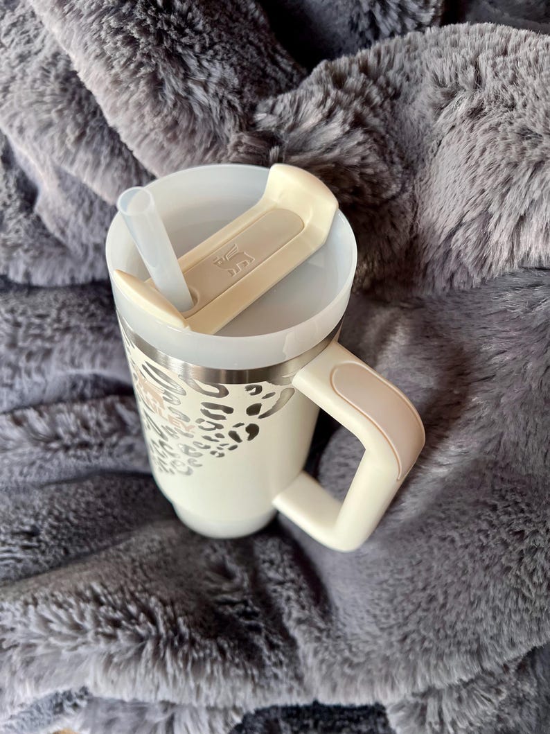 Puede incluir: Taza de viaje color crema con un dise&ntilde;o de estampado de leopardo plateado. La taza tiene un asa, una tapa con pajita y un cierre abatible color crema. La taza est&aacute; sobre una manta gris y esponjosa.