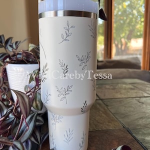 Pu&ograve; includere: Un bicchiere bianco con coperchio azzurro e cannuccia. Il bicchiere &egrave; decorato con un delicato motivo a foglie grigie. Il testo "With Careby Tessa" &egrave; visibile.