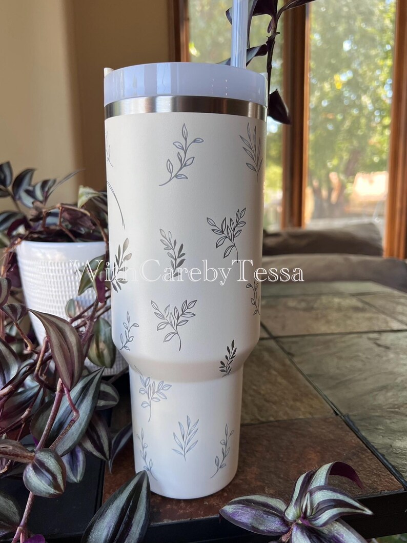 Pu&ograve; includere: Un bicchiere bianco con coperchio azzurro e cannuccia. Il bicchiere &egrave; decorato con un delicato motivo a foglie grigie. Il testo "With Careby Tessa" &egrave; visibile.