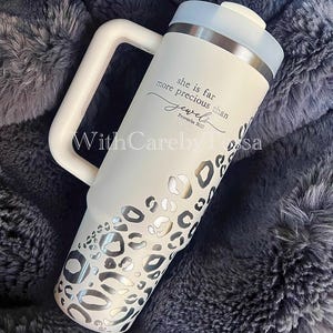 Puede incluir: Un vaso aislado blanco con asa, con un dise&ntilde;o de estampado de leopardo plateado y el texto "she is far more precious than jewels Proverbs 31:10". El vaso tiene una tapa blanca y un borde plateado.