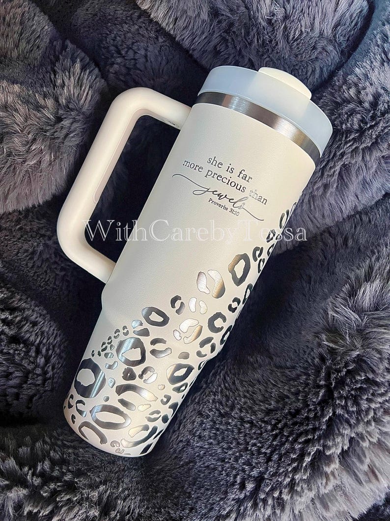 Puede incluir: Un vaso aislado blanco con asa, con un dise&ntilde;o de estampado de leopardo plateado y el texto "she is far more precious than jewels Proverbs 31:10". El vaso tiene una tapa blanca y un borde plateado.
