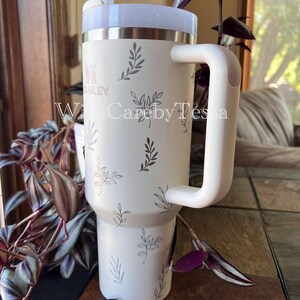 Pu&ograve; includere: Bicchiere Stanley color crema con manico e coperchio bianchi, decorato con un delicato motivo a foglie grigie. Il bicchiere include una cannuccia ed &egrave; progettato per bevande. Il testo "WithCarebyTessa" &egrave; visibile sul bicchiere.