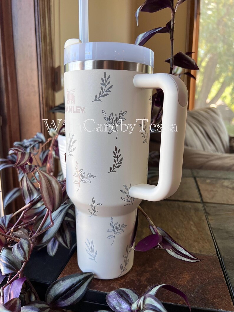 Pu&ograve; includere: Bicchiere Stanley color crema con manico e coperchio bianchi, decorato con un delicato motivo a foglie grigie. Il bicchiere include una cannuccia ed &egrave; progettato per bevande. Il testo "WithCarebyTessa" &egrave; visibile sul bicchiere.