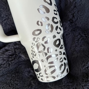 Puede incluir: Vaso blanco con asa, con un dise&ntilde;o de estampado de leopardo plateado. El vaso tiene forma curva y un asa blanca. El texto "Proverbios" es visible. El vaso est&aacute; sobre una superficie texturizada gris oscuro.