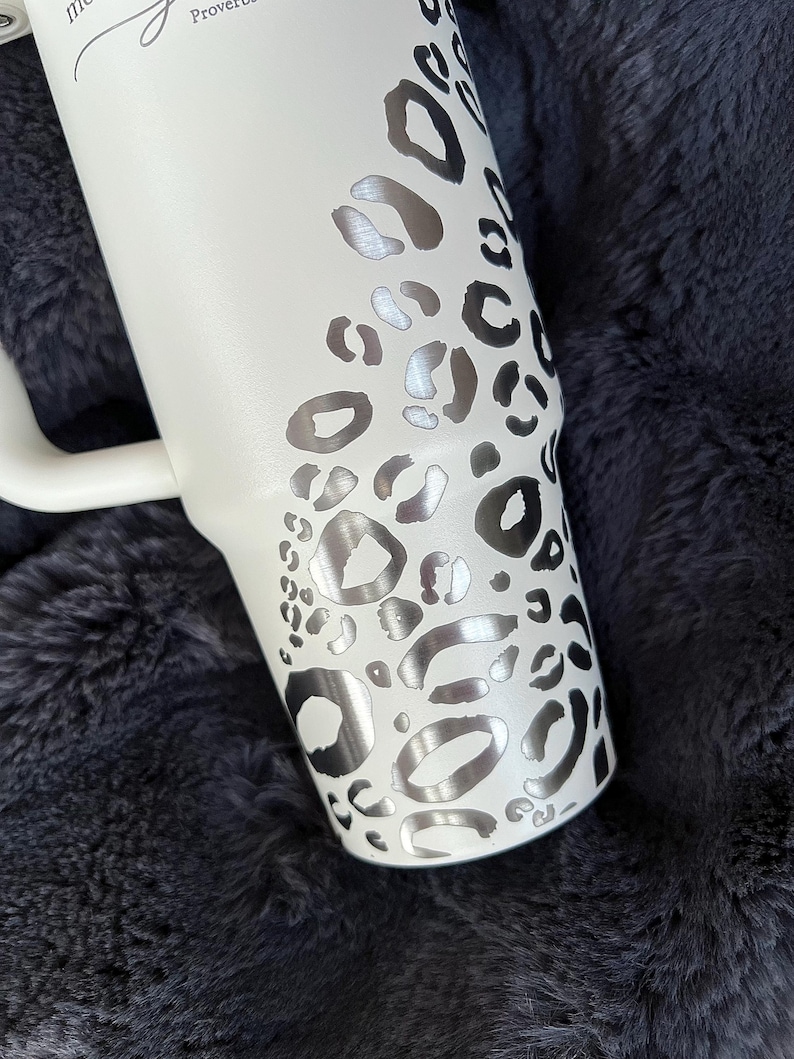 Puede incluir: Vaso blanco con asa, con un dise&ntilde;o de estampado de leopardo plateado. El vaso tiene forma curva y un asa blanca. El texto "Proverbios" es visible. El vaso est&aacute; sobre una superficie texturizada gris oscuro.