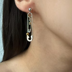 Può includere: Orecchini a spilla da balia argentati con un design pendente. Ogni orecchino presenta un piccolo perno a sfera argentato, un anello circolare e due spille da balia di diverse dimensioni. Piccole perline color oro aggiungono un tocco di colore.