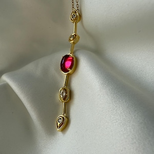 May include: A gold necklace with a vertical design. It features a large, oval, red gemstone and several smaller, clear gemstones. The necklace has a delicate chain and is displayed against a white, silky background.