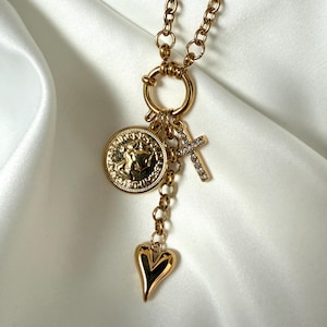 Puede incluir: Collar dorado con múltiples colgantes. Incluye un colgante de moneda, una cruz con pequeños cristales y un dije en forma de corazón. La cadena tiene un diseño de eslabones gruesos. El fondo es una tela blanca y sedosa.