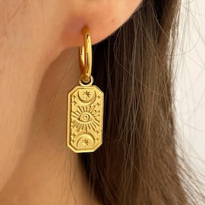 Peut inclure: Boucles d'oreilles créoles dorées avec un pendentif orné d'un motif céleste avec un soleil, une lune, des étoiles et un œil.