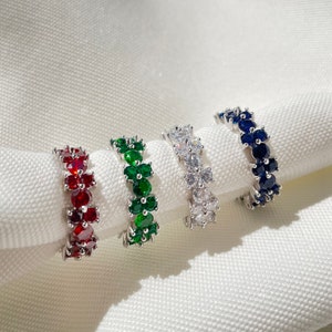 May include: Four silver rings featuring different coloured gemstones. Each ring is adorned with a row of round stones. The stones are red, green, white, and blue.