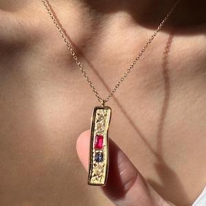 Może przedstawiać: Złoty naszyjnik z wisiorkiem w kształcie prostokąta. Wisiorek zawiera szereg kolorowych kamieni szlachetnych, w tym czerwone, niebieskie i przezroczyste kamienie. Naszyjnik jest noszony na szyi.