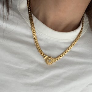 Könnte beinhalten: Eine goldene Kettenkette mit einem runden Anhänger, der einen klaren Stein in der Mitte trägt.