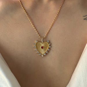 Puede incluir: Collar dorado con colgante en forma de corazón. El colgante presenta una piedra central roja, rodeada por un diseño de rayos de sol y cristales transparentes. El collar se lleva alrededor del cuello.