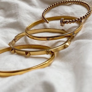May include: Four gold bracelets stacked on top of each other. The top bracelet has a beaded design and a small gold charm with a clear stone. The second bracelet has a plain band with clear stones. The third bracelet has a plain band with small gold spikes. The bottom bracelet has a plain band with the word "BEAUSANCY" engraved on it.