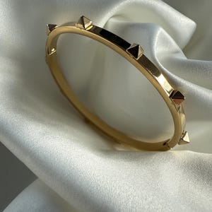Könnte beinhalten: Goldfarbenes Armband mit pyramidenförmigen Nieten. Das Armband ist im Bangle-Stil gehalten und hat eine glatte, polierte Oberfläche. Die Nieten sind gleichmäßig um das Band angeordnet. Das Armband wird auf einem weißen, seidigen Stoff präsentiert.
