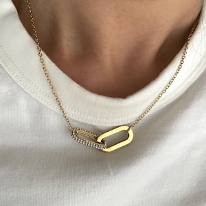 Op de afbeelding: Goudkleurige ketting met twee in elkaar grijpende schakels. Eén schakel is rechthoekig en effen, de andere is een kleinere, rechthoekige schakel bezet met kleine, heldere kristallen. De ketting wordt gedragen tegen een witte achtergrond.