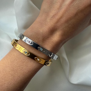 Op de afbeelding: Twee metalen armbanden, één goudkleurig en één zilverkleurig, worden getoond. Elke armband is versierd met kleine, kleurrijke oogvormige details. De goudkleurige armband heeft blauwe, groene en heldere accenten, terwijl de zilverkleurige armband roze, blauwe en heldere accenten heeft.