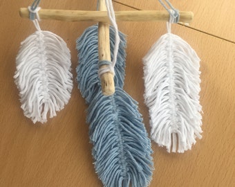 Puede incluir: Un tapiz de macramé de pared con tres plumas. Las plumas están hechas de hilo blanco y azul claro y están unidas a una varilla de madera. La varilla está suspendida del techo con cuerda.