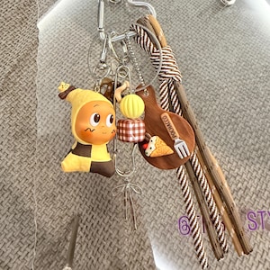 Puede incluir: Una colección de llaveros con un personaje de dibujos animados en amarillo y negro, una guitarra de madera con un taco y cuentas decorativas. Los llaveros están sujetos a un cierre plateado en forma de corazón y un cordón trenzado marrón y beige.