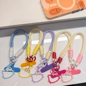 May include: A collection of colorful phone charms featuring a cartoon cat design. Each charm has a different color scheme, including blue, yellow, purple, and pink. The charms are attached to a lanyard-style strap.