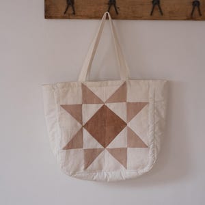 May include: A quilted tote bag featuring a central star pattern composed of cream, beige, and brown fabric patches. The bag has two long fabric handles and a soft, padded texture throughout, showcasing a geometric design on the front panel.