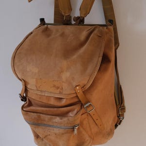 May include: A vintage brown leather backpack with a flap closure and multiple pockets. The backpack has adjustable straps and a zipper pocket on the front. It is hanging on a wooden hanger.