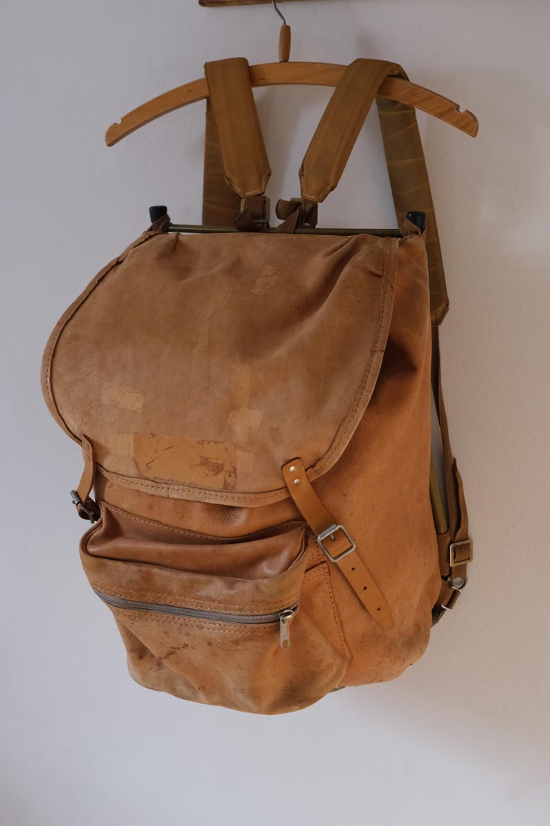 May include: A vintage brown leather backpack with a flap closure and multiple pockets. The backpack has adjustable straps and a zipper pocket on the front. It is hanging on a wooden hanger.