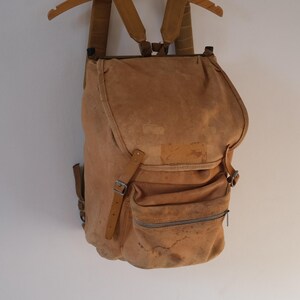 May include: A tan suede backpack with adjustable straps and multiple pockets. The vintage-style bag has a flap closure and a zippered front pocket. The backpack is hanging on a wooden hanger against a white background.