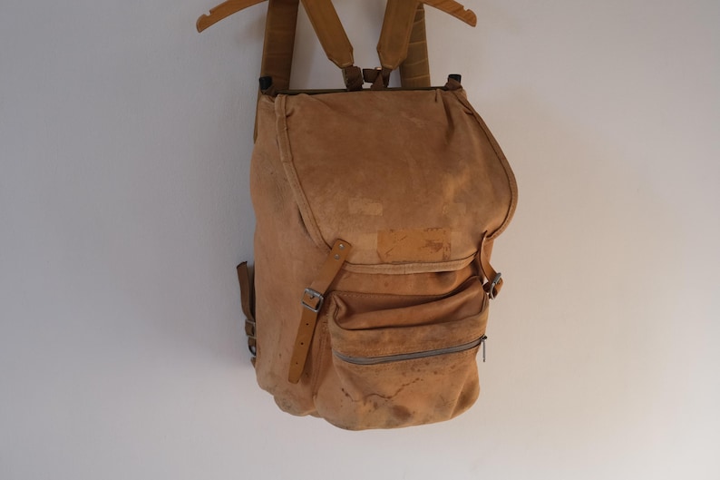 May include: A tan suede backpack with adjustable straps and multiple pockets. The vintage-style bag has a flap closure and a zippered front pocket. The backpack is hanging on a wooden hanger against a white background.