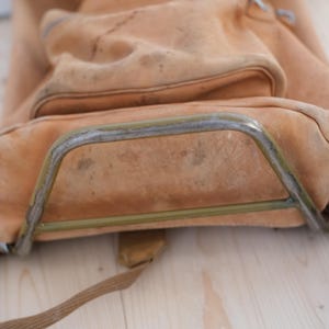 May include: A worn, tan leather backpack with a metal frame. The bag has a large main compartment and a smaller front pocket. The leather shows signs of wear and tear, with visible stains and discoloration. The metal frame is slightly rusted.