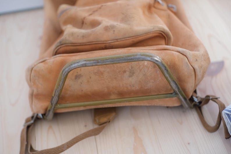 May include: A worn, tan leather backpack with a metal frame. The bag has a large main compartment and a smaller front pocket. The leather shows signs of wear and tear, with visible stains and discoloration. The metal frame is slightly rusted.