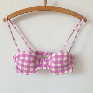 May include: A pink and white gingham bikini top with adjustable straps, hanging on a wooden hanger. The bikini top has a classic underwire design and a clasp closure at the back. The fabric is a light, summery material.