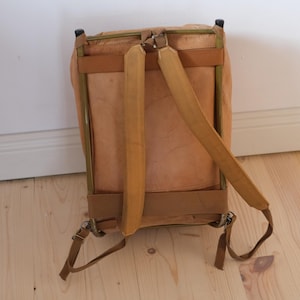 May include: A vintage-style backpack with a light brown leather exterior and tan straps. The backpack has a rectangular shape with a metal frame and adjustable shoulder straps. The bag is set against a light wood floor and white wall.