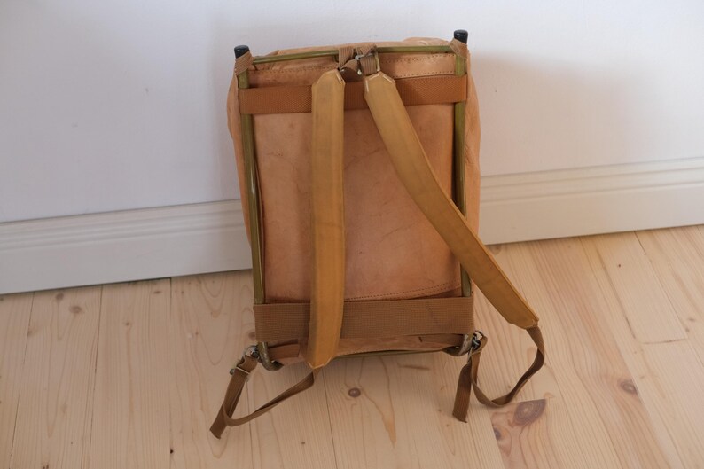 May include: A vintage-style backpack with a light brown leather exterior and tan straps. The backpack has a rectangular shape with a metal frame and adjustable shoulder straps. The bag is set against a light wood floor and white wall.