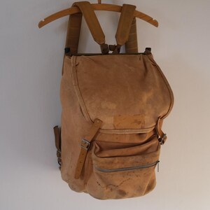 May include: A tan canvas backpack with a flap closure and multiple pockets. The backpack has adjustable shoulder straps and is suspended from a wooden hanger. The bag appears worn, with visible stains and wear.
