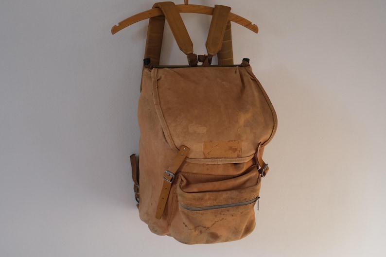 May include: A tan canvas backpack with a flap closure and multiple pockets. The backpack has adjustable shoulder straps and is suspended from a wooden hanger. The bag appears worn, with visible stains and wear.