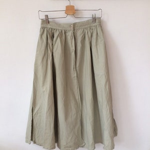 May include: A sage green midi skirt featuring a gathered waistband with a single centered button closure. The skirt has a relaxed, flowing silhouette with a vertical seam down the front and a side slit, hanging from a metal clothes hanger.