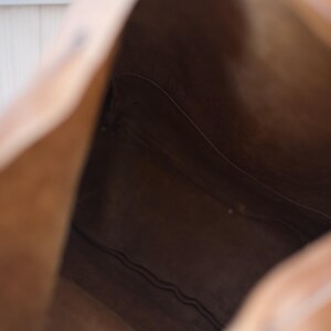 May include: A light brown leather tote bag, viewed from the inside. The bag's interior is a darker shade of brown, with visible seams and stitching. The bag's opening is visible, showing the depth and structure of the bag.
