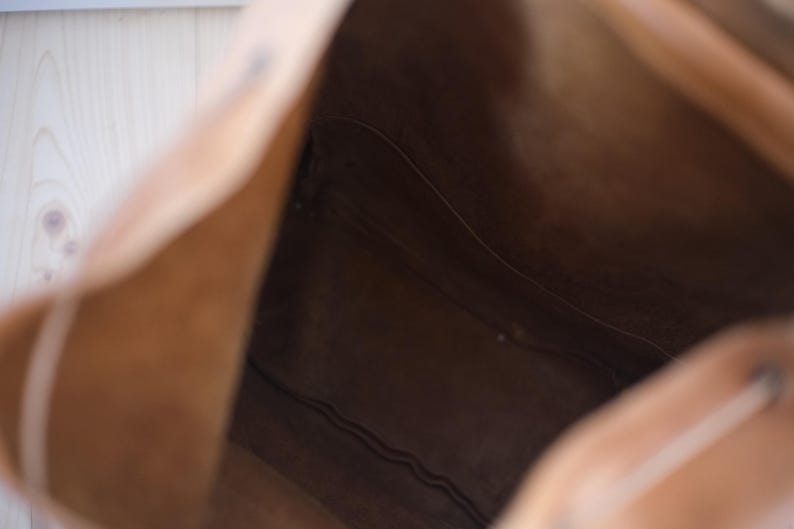 May include: A light brown leather tote bag, viewed from the inside. The bag's interior is a darker shade of brown, with visible seams and stitching. The bag's opening is visible, showing the depth and structure of the bag.