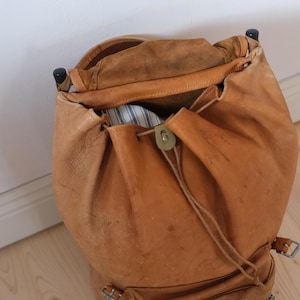 May include: A vintage, open leather backpack in a warm tan color. The bag features a drawstring closure with a brass clasp and adjustable straps. The interior reveals a glimpse of striped fabric. The backpack is designed with a classic, rustic aesthetic.